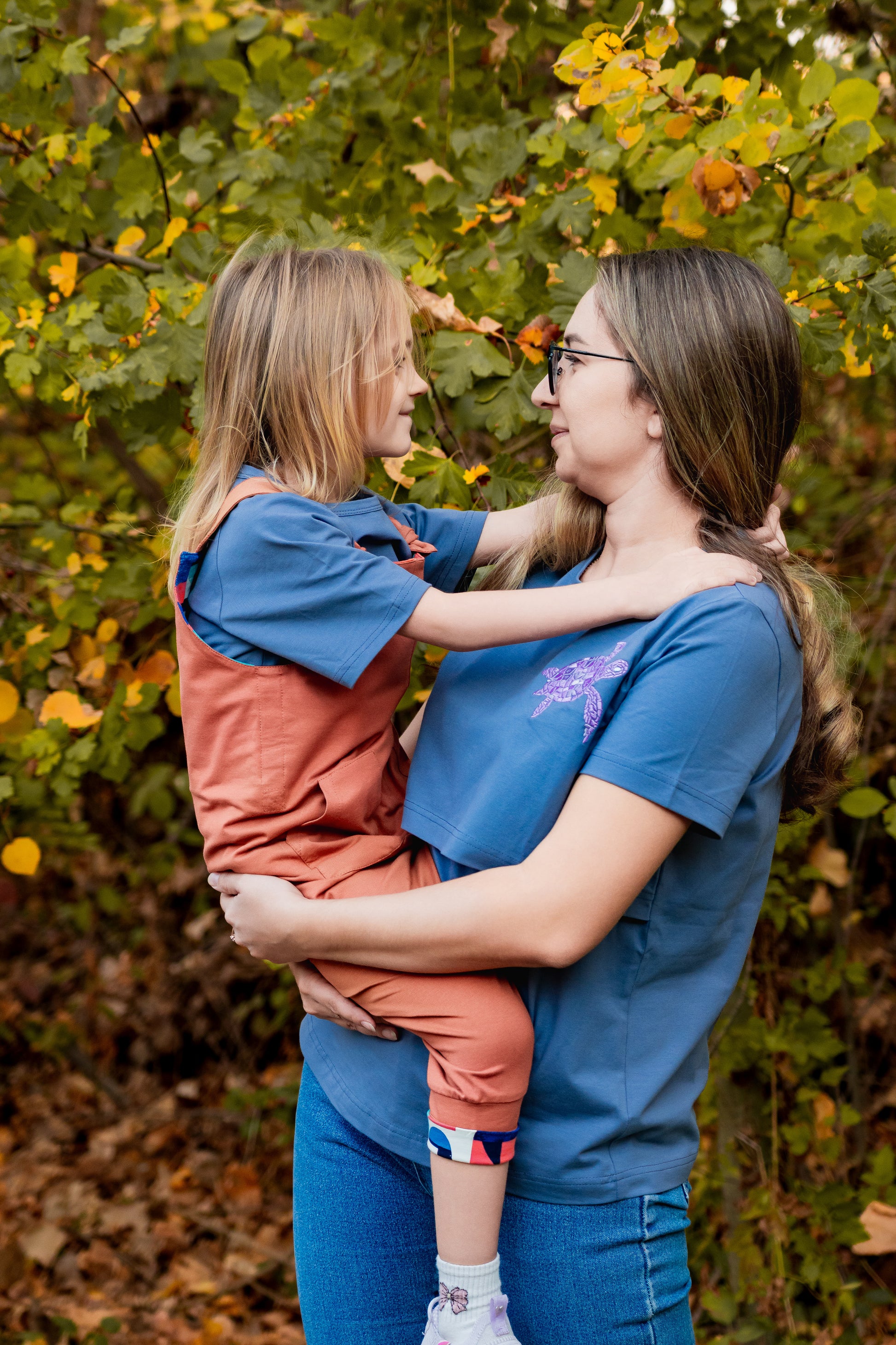 Woman holding a child in an outdoor setting with trees and leaves.